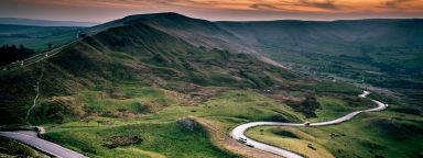 The winding road at Mam Tor. Mam Tor is green and grassy and the sky is red as if the sun is just starting to set.