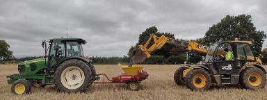 An image showing a front loader putting crushed basalt into a spreader behind a tractor, in a crop field.