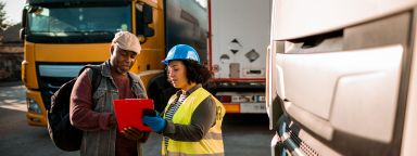 Woman in high-vis vest and hard hat shows clipboard to worker in front of lorries