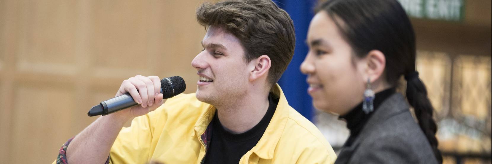 Two music students rehearsing - one is singing into a microphone, the other sites closer to the camera and plays the guitar