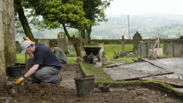 An archaeology student excavating soil at a dig site by a church.