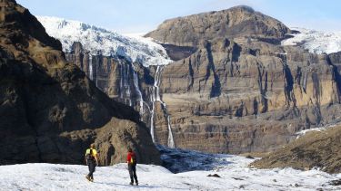 Two researchers stand on snow looking up at rock face