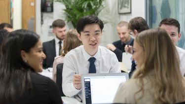 Three students are sitting and talking to each other while a laptop with their work is open on the table. Other students can be seen working in the background.
