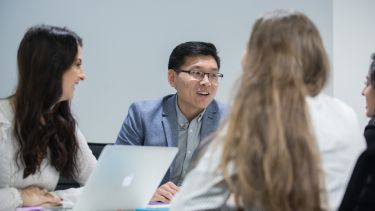 Four students sit around a table talking. A laptop is open on the table.