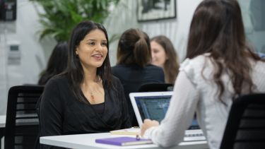 Two students talk while sitting and facing each other. A laptop is open between them. Other studenst can been seen in the background.