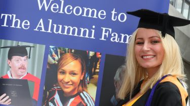 A female graduate stands in front of the alumni banner stand.
