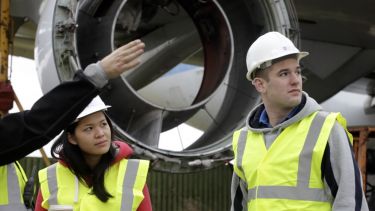 Two Civil Engineering students on a Year in Industry placement. They have on hi-vis vests and hard hats and are standing in front of a propeller.