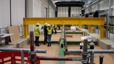 The yellow steel crossbeam and metallic structures of a large scale facility surround a group of researchers in high viz jackets and hardhats stood on sand