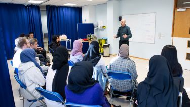 A group of students learn clinical skills from a lecturer. He is presenting to them. They are sitting down.