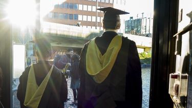 Two graduates walk through a doorway. The sun is reflecting on the glass of the door.