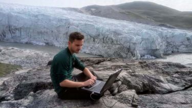 Alumnus Olly Bartlett sitting with a laptop in front of an ice sheet