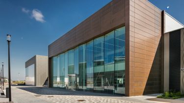 A bronze-coloured, modern, cuboid-shaped building with large glass-panelled front, with a modern and clean-looking concourse at the front