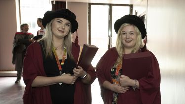 Two staff volunteers. They wear red robes, hats, lanyards and carry folders in their arms.
