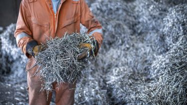 A person in orange protective clothing and gloves holding a handful of metal machining waste, standing in front of piles of machining waste