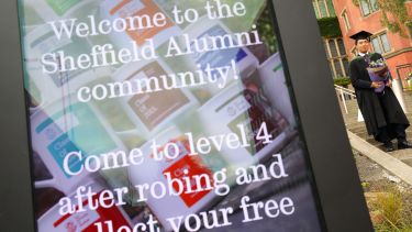 Sign for the Alumni Relations stand with a graduate in the background