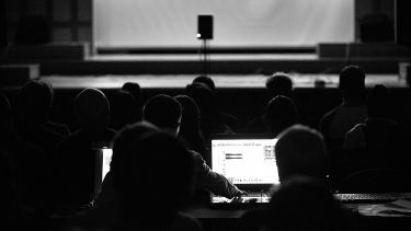 crowd sat looking at a stage, a person sat at the back of the crowd on a laptop.
