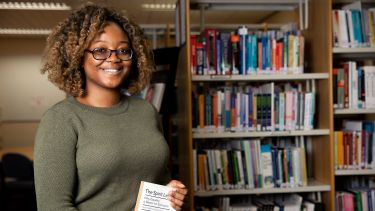 A female student poses in the School of Health and Related Research library with a book.