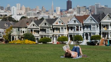 A woman reads a book on a grassy area. The city of San Francisco can be seen in the background. 