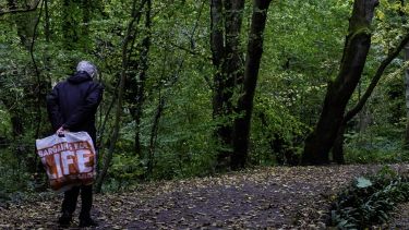 A man with a 'Bargains for Life' bag walks through the woods. 