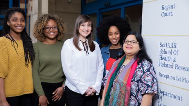 Five female students stand outside the School of Health and Related Research main entrance, next to the building's sign.