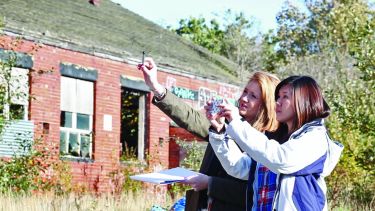 Students standing and looking at something off camera in front of abandoned overgrown building