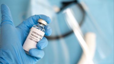 Picture of a hand in a blue glove holding up a small vaccine sample in a glass bottle