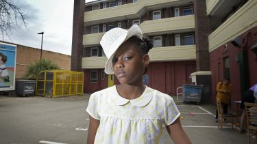 A young girl wearing a white had and top stands in front of an apartment building and looks at the camera 