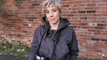 Philosophy alumna and photographer Laura Page stood in front of exposed brick wall with her camera
