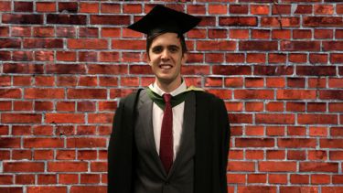 Graduate in cap and gown stands against wall