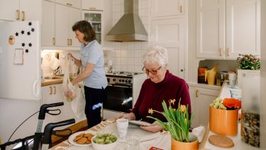 Senior woman using digital tablet while female healthcare worker holding grocery bag in kitchen