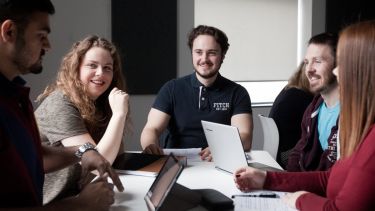 5 students sitting around a desk in a seminar room, talking and working on laptops