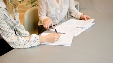 Woman signing on white paper, sitting beside another women organising paperwork