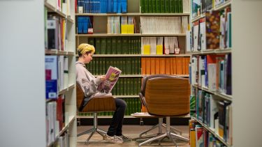 Student sat in chair reading book in library, surrounded by book shelves