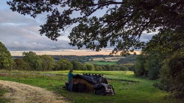 Green farmland and fields stretch out in the distance. In the foreground a man is covering some hay bales with black plastic