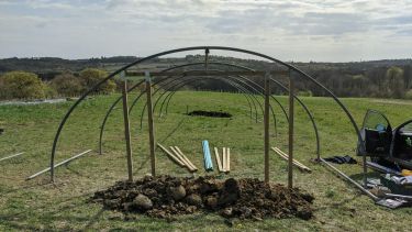 A green field with the frame of a polytunnel build on it. Under the frame are a set of wooden poles and the frame covering.