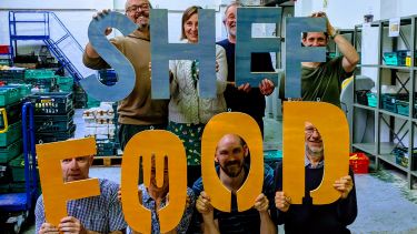 Photo of a group of eight adults in a warehouse holding up a letter each. The letters read SHEFFOOD.