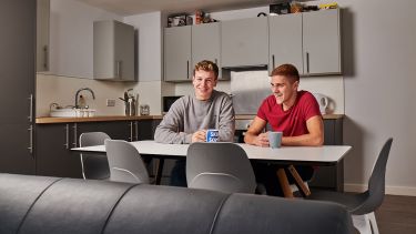 two male students sat at a dining table in a kitchen with mugs