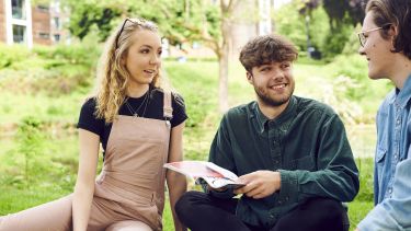 three students sat together on the grass with a magazine