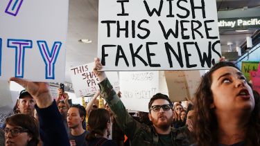 Protesters at an event, one holding a placard reading "I wish this were fake news"