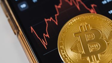 A coin with a Bitcoin symbol lying on a mobile phone screen with an image of stock markets