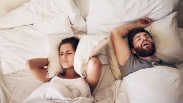 A young couple in bed with the man lying on his back snoring and the woman covering her ears with a pillow