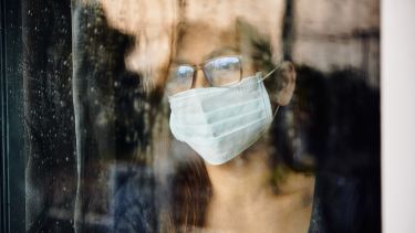 A woman behind a window pane with face mask