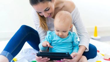 Baby sat between woman's arms, playing on tablet the woman is holding