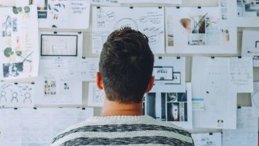 A photograph of a man looking at a noticeboard. On the noticeboard are sheets of paper showing sketches, notes, images and graphs.