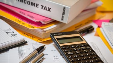 The image include pens, documents detailing tax information, a calculator, a box called Income Tax and a diary.