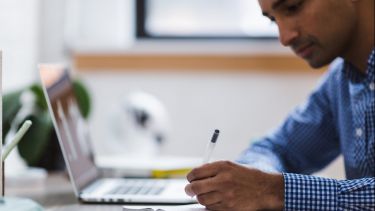 A photograph of an Asian man writing in a notebook. Beside him on his desk is a laptop.