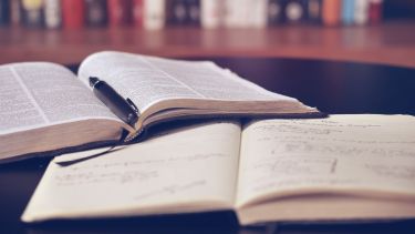 An image of an open printed book with a pen laid on it. Also on the table is a notebook with notes written into it.