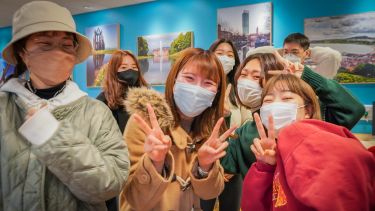 A group of 7 happy students wearing facemasks and making the v sign with their fingers in the Oasis Cafe at the English Language Teaching Centre