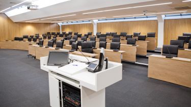 A photograph of the Middleton Lecture Theatre. The presenter's desk is in the foreground. In the background a rows of wooden lecture theatre desks with leather chairs.