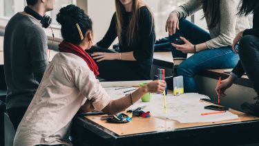 A group of people sat round a table working together. On the table is stationery and papers.